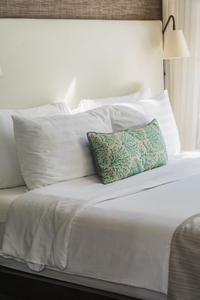 A neatly made bed with white linens, two white pillows, and a decorative pillow with a green coral pattern, positioned next to a wall lamp.