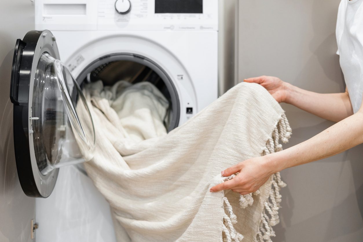 A person is removing a light-colored blanket with tassels from a front-loading washing machine.
