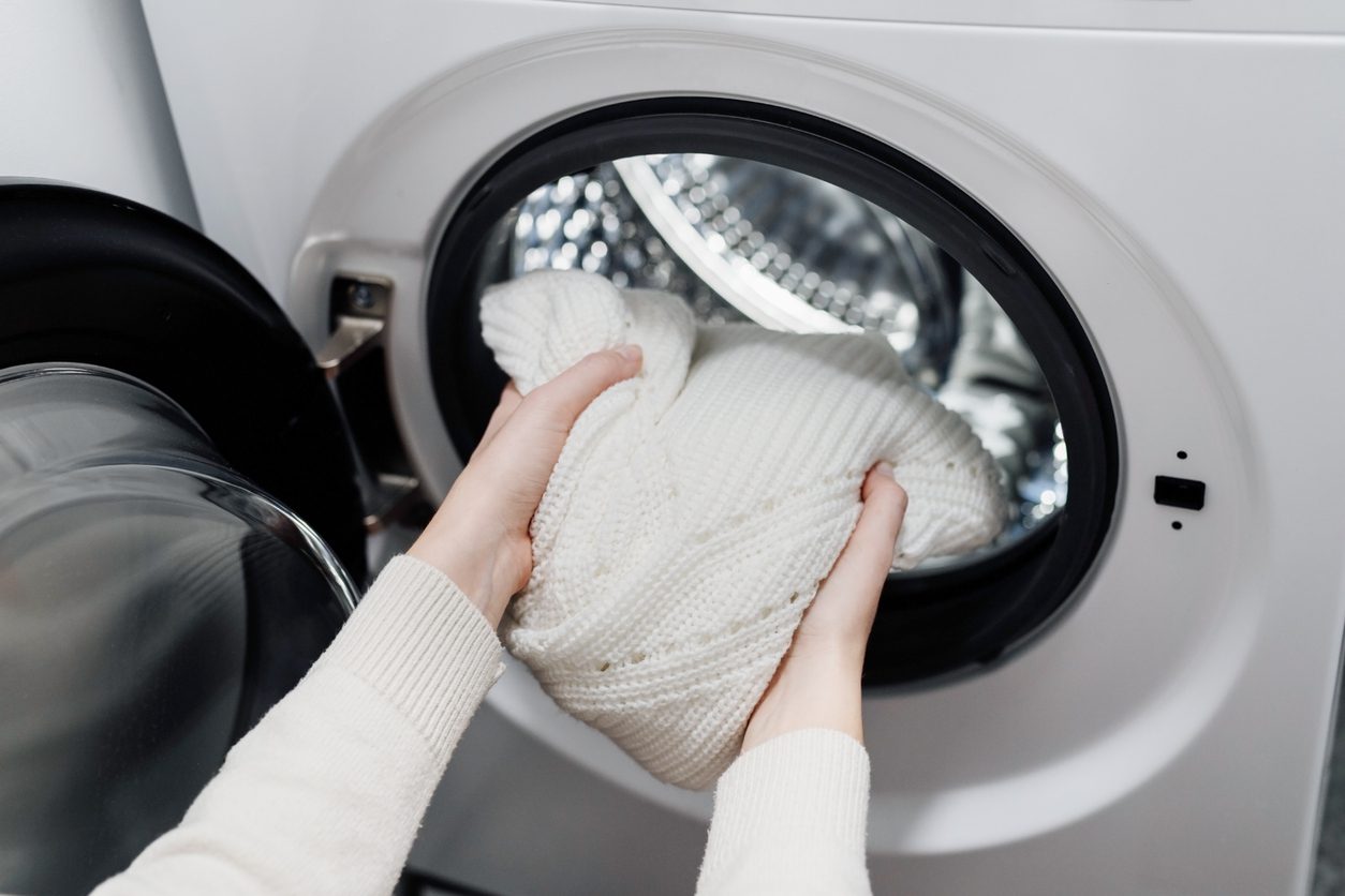Person placing a white knitted sweater into the drum of a front-loading washing machine.