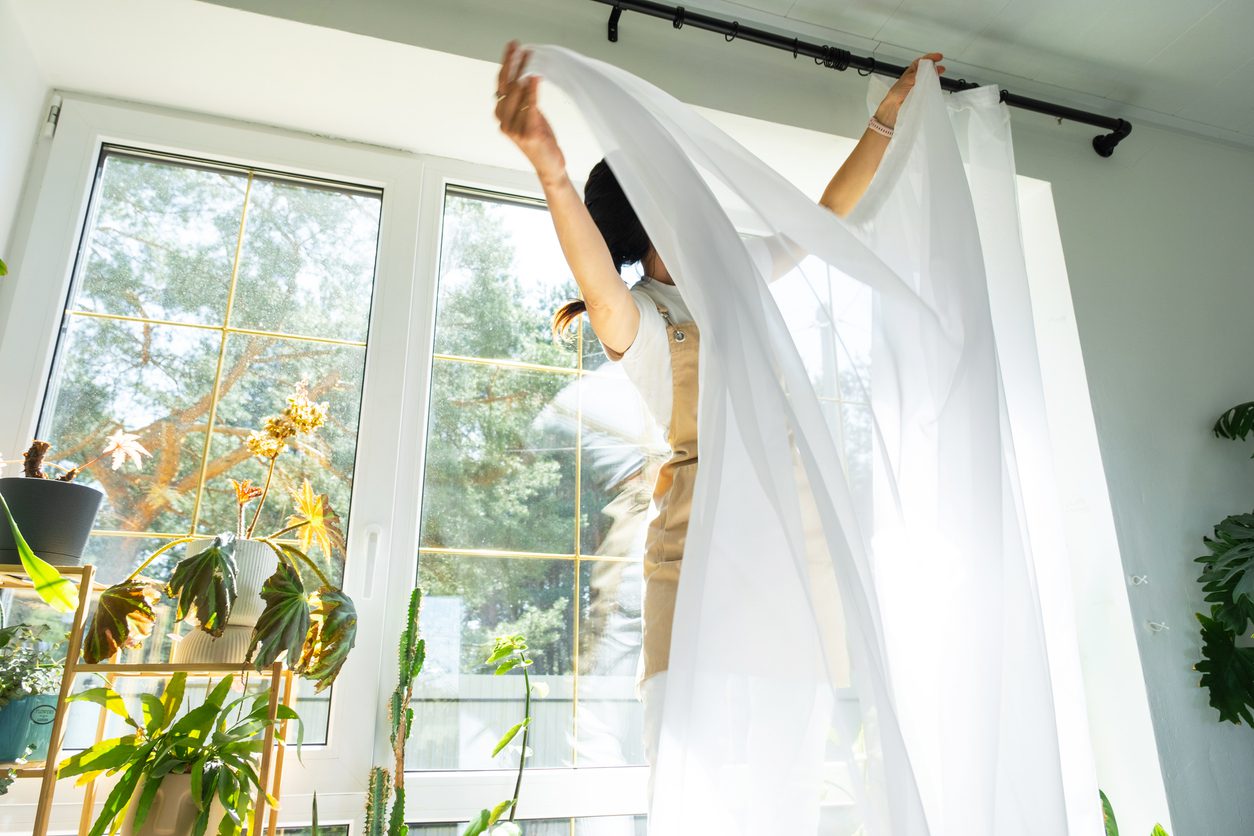Person hanging white curtains on a rod in front of a large window, surrounded by houseplants in a bright, sunlit room.