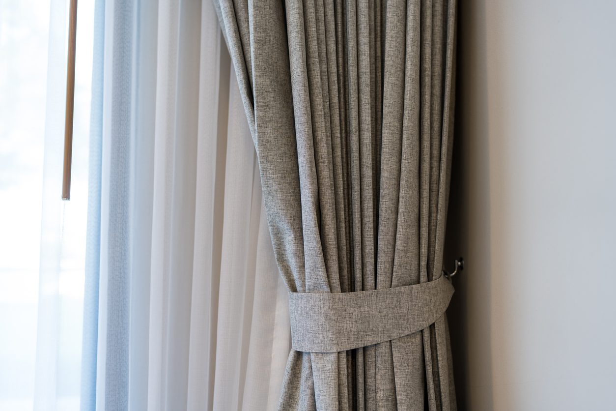 Close-up of a grey curtain tied back with a matching fabric tie, next to a sheer white curtain and a white wall.